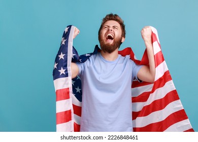 Portrait of positive overjoyed bearded man holing huge american flag and rejoicing while celebrating national holiday, looking up and yelling. Indoor studio shot isolated on blue background. - Powered by Shutterstock