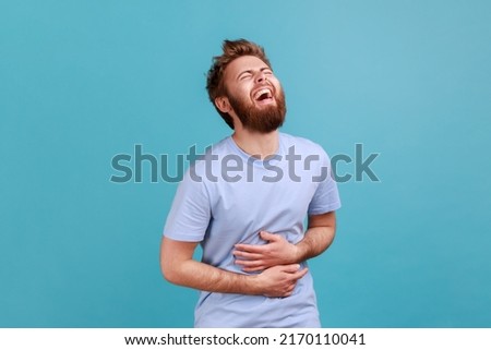 Portrait of positive overjoyed bearded man laughing happily at something keeps hands on belly, smiles broadly, expressing positive emotions. Indoor studio shot isolated on blue background.