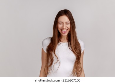 Portrait Of Positive Playful Woman Standing With Closed Eyes And Tongue Out, Demonstrating Childish Behavior, Wearing White T-shirt. Indoor Studio Shot Isolated On Gray Background.