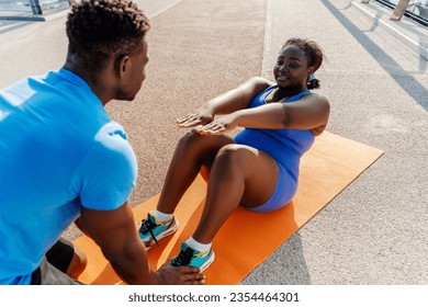 Portrait of positive plus size African woman training with personal trainer, do abs, sitting on yoga mat outdoors. Sports motivation, healthy lifestyle, body transformation - Powered by Shutterstock