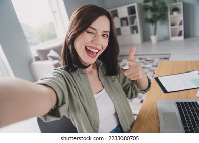 Portrait of positive pretty girl sit chair take selfie record video eye wink pointing finger you workplace indoors