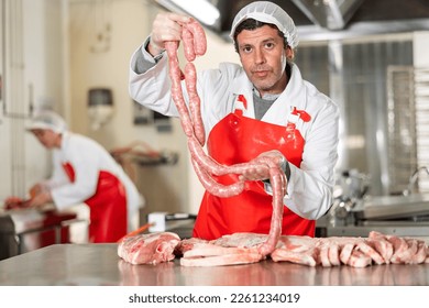 Portrait of positive professional butcher in white uniform and red apron holding chain of raw sausages while standing in processing workshop near fresh meat cutting table - Powered by Shutterstock