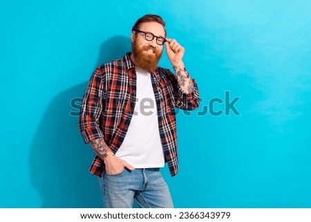 Portrait of positive satisfied man with tattoo wear plaid shirt touching eyewear hold arm in pocket isolated on blue color background