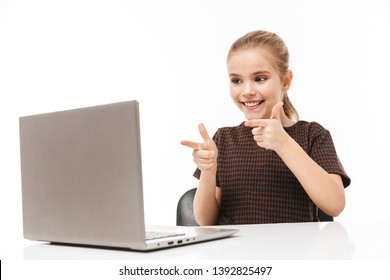 Portrait Of Positive School Girl Rejoicing And Using Silver Laptop While Sitting At Desk In Class Isolated Over White Background