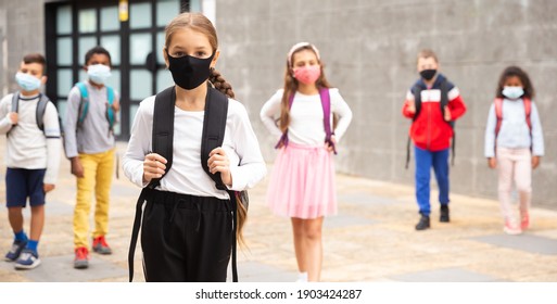 Portrait Of Positive Schoolgirl In Mask Standing Near School, Children On Background
