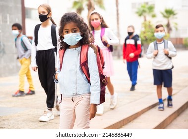 Portrait Of Positive Schoolgirl In Mask Standing Near School, Children On Background