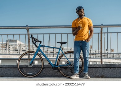 Portrait of positive smiling African man wearing safety helmet holding mobile phone standing near bicycle on street. Handsome rider checking email, online shopping. Technology concept - Powered by Shutterstock