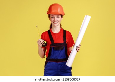 Portrait of positive woman builder holding blueprints and roulette for measuring, looking at camera with smile, wearing overalls and protective helmet. Indoor studio shot isolated on yellow background
