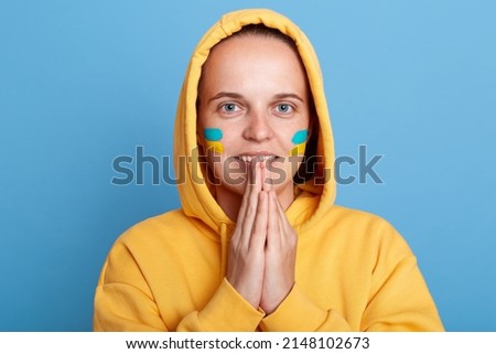 Portrait of positive woman in yellow hoodie with Ukrainian flag on cheeks, keeps palms together, thanks God for ending war in Ukraine, posing isolated over blue background.