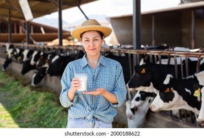 Portrait Of Positive Young Adult Woman Farmer With Glass Of Fresh Milk Working In Cowshed At Dairy Farm