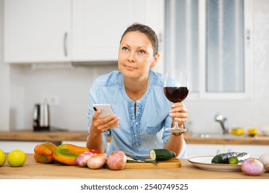 Portrait of positive young adult woman holding glass of wine and reading message on her smartphone while standing at kitchen interior near countertop with fresh vegetables - Powered by Shutterstock