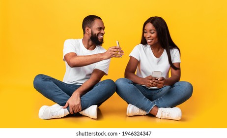 Portrait of positive young African American couple sitting on the floor, using smartphones. Smiling bearded man showing his gadget to woman, sharing social media content, yellow studio wall