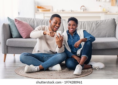 Portrait of positive young African American couple sitting on the floor carpet, using smartphone. Smiling man holding his gadget and pointing at screen, showing photos, sharing social media content - Powered by Shutterstock