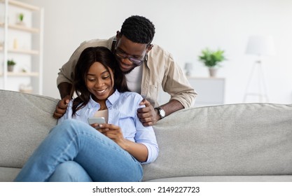 Portrait of positive young African American couple sitting on the couch, using smartphone. Smiling woman holding her gadget and sharing social media content, showing photos or videos to her husband - Powered by Shutterstock