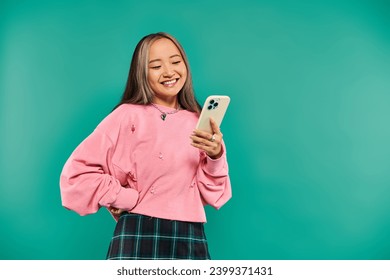 portrait of positive young asian woman in pink sweatshirt and plaid skirt using smartphone on blue - Powered by Shutterstock