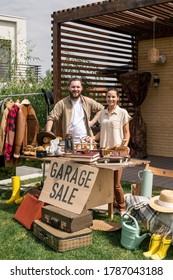 Portrait Of Positive Young Couple Standing At Table With Useless Things And Selling It At Garage Sale In Backyard