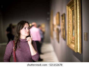 Portrait of positive young girl attentively looking at paintings in art museum 
