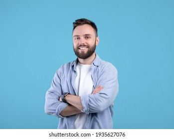 Portrait of positive young guy posing with crossed arms and smiling at camera on blue studio background