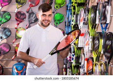 Portrait Of Positive Young Man Looking For Tennis Racquet In Store Of Sports Equipment