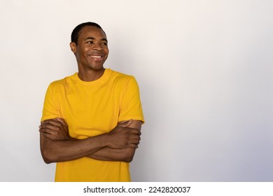 Portrait of positive young man posing with folded arms against white background. African American guy wearing yellow T-shirt looking away and smiling. Happiness concept - Shutterstock ID 2242820037
