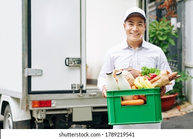Portrait of positive young Vietnamese courier holding plastic box with fresh fruits, bread and bottled milk