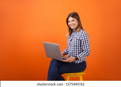 Portrait Of Positive Young Woman With Brown Hair In Checkered Casual Shirt And Denim Sitting With Toothy Smile, Working On Laptop, Looking At Camera. Indoor Studio Shot Isolated On Orange Background