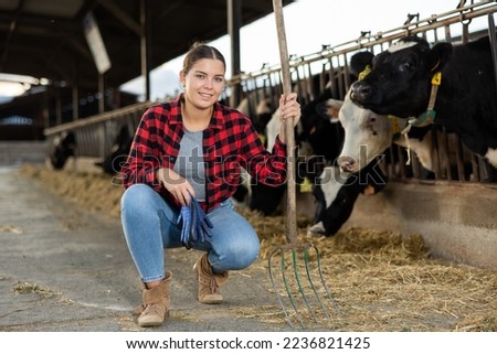 Portrait of positive young woman farmer with pitchfork squatting in cowshed among rows of cows in stalls.