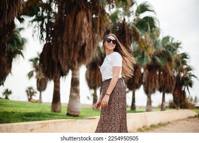 Portrait of positive young woman wearing summer clothes and sunglasses standing on hotel territory with palm trees around. Charming lady smiling and looking at camera. Winter at Cyprus - Shutterstock ID 2254950505