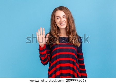 Portrait of positive young woman wearing striped casual style sweater standing waving hand, looking at camera with engaging toothy smile. Indoor studio shot isolated on blue background.