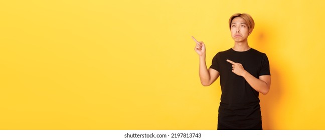 Portrait Of Pouting Upset And Funny Asian Guy Pointing And Looking Upper Left Corner With Regret, Standing Yellow Background