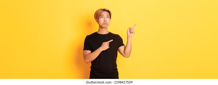 Portrait Of Pouting Upset And Funny Asian Guy Pointing And Looking Upper Left Corner With Regret, Standing Yellow Background