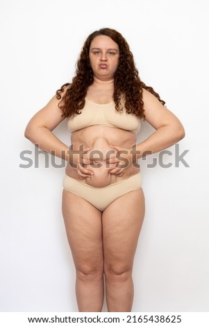 Portrait of pouty overweight woman with long curly hair wearing beige underwear, squeezing excess fat of belly, looking offended, posing on white background. Weight loss, obesity, health problems.