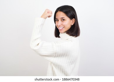 Portrait Of Powerful Cheerful Young Brunette Woman Wearing White Knitted Sweater Against White Background Showing Muscles.