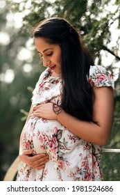 Portrait Of Pregnant Woman With Long Black Hair In Park Dressed In White Dress With Flowers, Woman Holds Her Baby Belly, Pregnant Woman In Stands In Park, Indian Woman Portrait