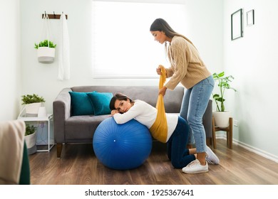 Portrait Of A Pregnant Woman On Her Knees And Resting Her Head On A Fitnes Ball At Home While Her Midwife Holds A Rebozo Around Her Belly