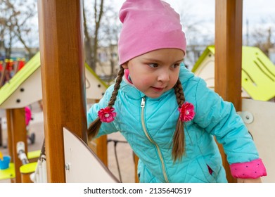 Portrait Of A Preschool Child On The Playground. A Five-year-old Girl In A Pink Hat Walks Up And Down The Stairs Holding Onto The Railing. Childhood. View From The Bottom Up.