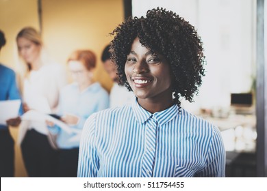 Portrait Of Pretty African American Business Woman With Afro Smiling At The Camera.Coworking Team On Background In Modern Office. Horizontal,blurred