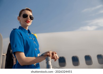 Portrait of pretty air stewardess in blue uniform and sunglasses looking away, standing on airstair on a daytime. Aircrew, occupation concept - Shutterstock ID 2026324451