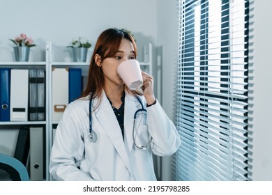 Portrait Of Pretty Asian Female Medical Worker With A Stethoscope Is Drinking Coffee While Taking A Break By The Window In A Bright Hospital Office At Daytime.