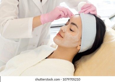 Portrait Of Pretty Asian Woman Enjoying Beauty Treatment In Spa With Cosmetologist  Performing Facial Care Procedures