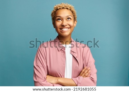 Portrait of pretty black girl of 20s looking up smiling biting lips, planning something, remembering funny moments, thinking about her boyfriend, standing with crossed hands on blue background