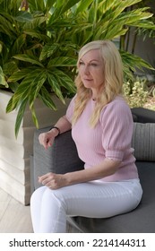 Portrait Of The Pretty Blond Haired Woman In Pink Blouse And White Pants, With Blue Eyes, Sitting On The Gray Couch, Not Looking Into The Camera, Smiling