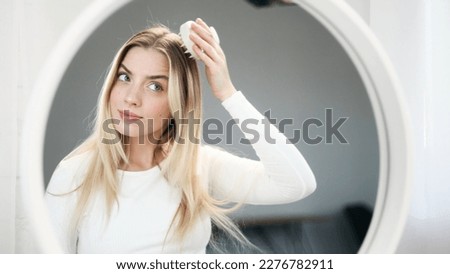 Portrait of pretty blond woman looking at mirror reflection and doing self hair scalp massage with massager at home Concept of hair loss growth stimulation trichology self care