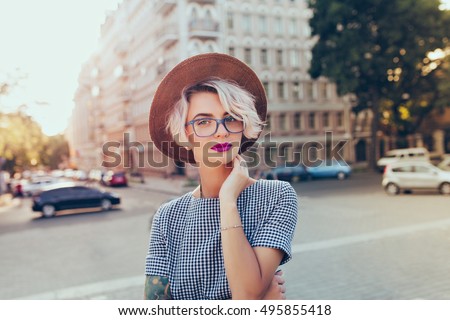 Portrait of pretty blonde girl with short hair posing to the camera on the street in city. She wears gray checkered  dress, glasses, hat and has purple lips.