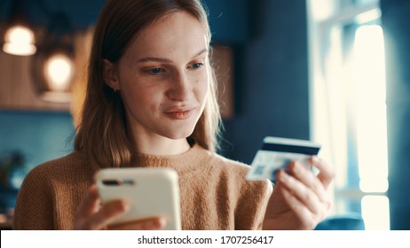 Portrait of pretty blondehair girl sitting on sofa in modern room, apartment and makes online payments through the Internet from bank card on smartphone. Online banking with smart phone