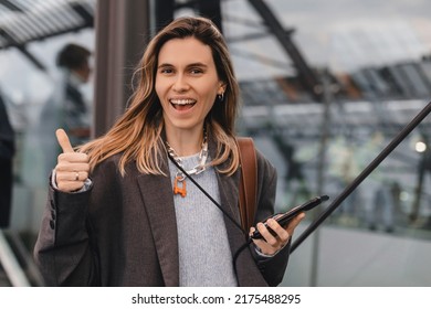 Portrait Of A Pretty Casual Girl Holding Mobile Phone And Showing Thumbs Up Standing On Bus Station. Blonde Woman Wear Grey Jacket, Brown Bag And Necklace. Good News, Win, Glory.