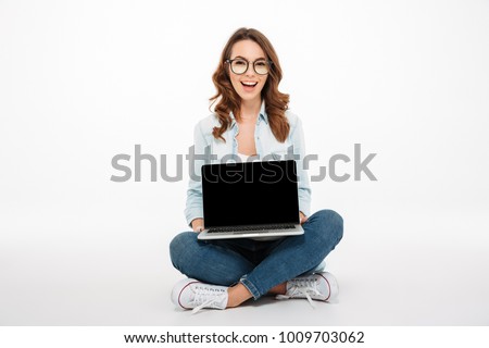 Portrait of a pretty casual girl showing blank screen laptop computer while sitting on a floor isolated over white background
