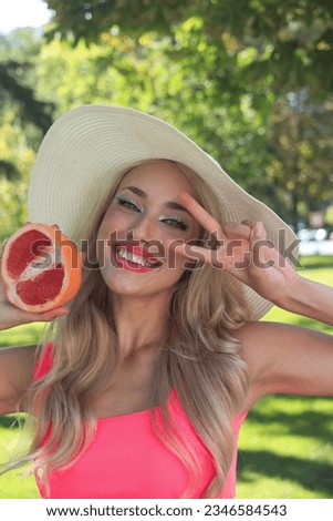 Portrait pretty caucasian blonde woman, smiling at the camera. She is holding a grapefruit in front of her eye. Healthy lifestyle. Barbie makeup.
