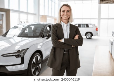 Portrait of pretty Caucasian vehicle saleswoman in suit standing with arms crossed near new modern car in bright and spacious showroom. Professional female salesperson at car dealership. - Powered by Shutterstock