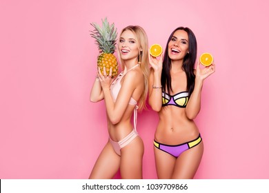 Portrait of pretty, charming, laughing, stylish trendy ladies, tourists with modern hairstyle in swimsuit showing ananas and orange in hands, standing over pink background, brunette vs blonde